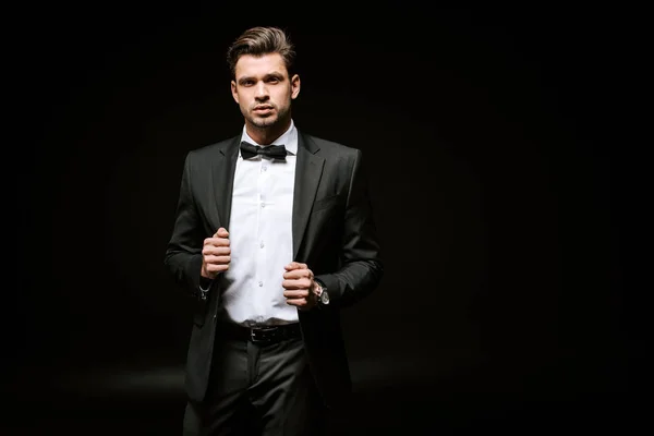 Uomo elegante in giacca e cravatta guardando la fotocamera e toccando blazer su nero — Foto stock