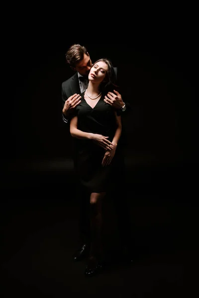 Мужчина стоит и целует красивую женщину в платье на черном — стоковое фото