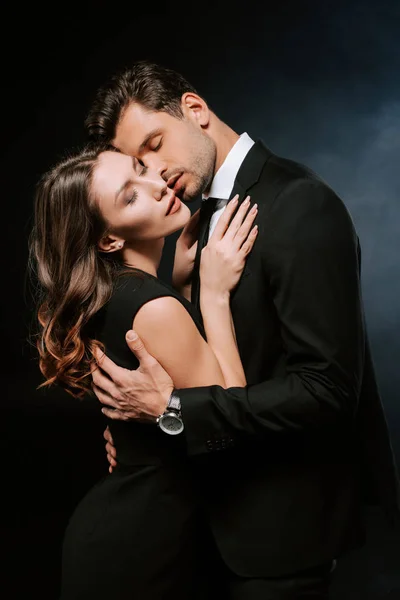 Guapo hombre abrazando atractiva mujer en negro con humo - foto de stock