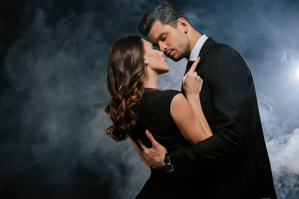 Atractiva mujer abrazando guapo hombre en negro con humo - foto de stock