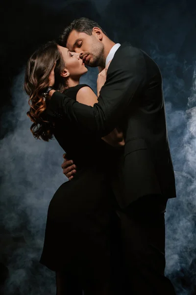 Hombre guapo en traje abrazando sensual mujer en negro con humo - foto de stock