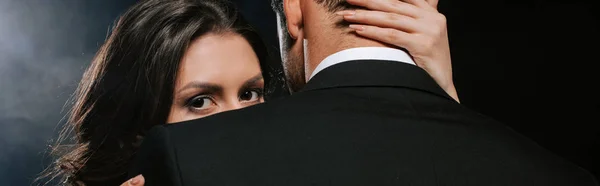 Panoramic shot of woman hugging man and looking at camera on black — Stock Photo