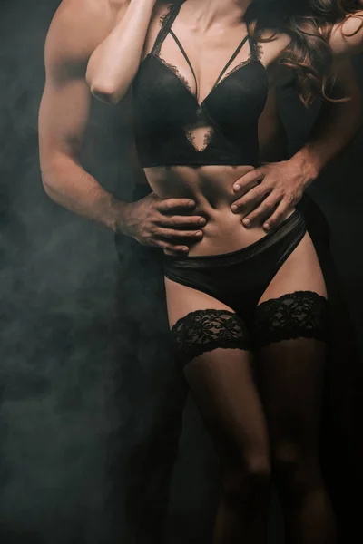 Corte vista de homem apaixonado abraçando jovem mulher em roupa interior em preto com fumaça — Fotografia de Stock