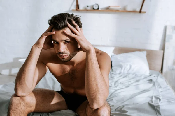 Homem sem camisa olhando para a câmera enquanto sentado na cama e tocando a cabeça — Fotografia de Stock