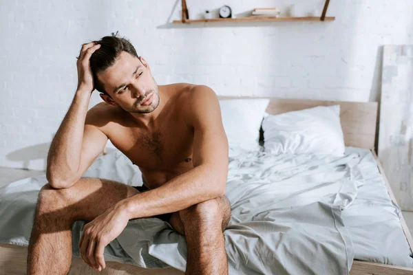 Uomo muscoloso guardando lontano mentre seduto sul letto e toccando i capelli — Foto stock