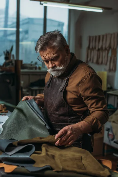 Atento, zapatero senior mirando piezas de cuero genuino en el taller - foto de stock