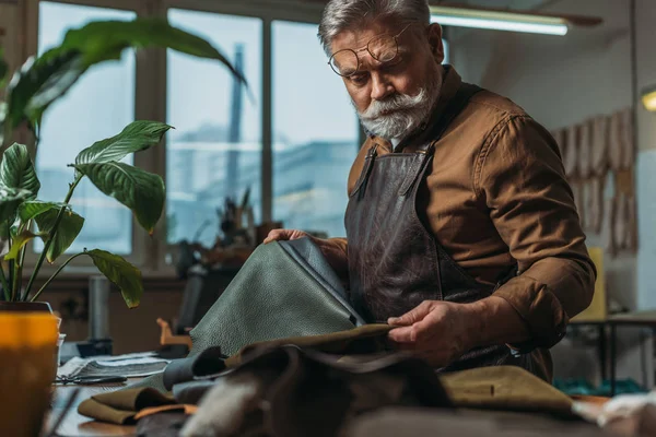 Selective focus of senior cobbler holding pieces of genuine leather in workshop — Stock Photo