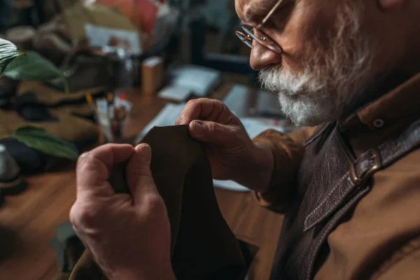 Attentive, senior shoemaker holding piece of genuine leather in workshop — Stock Photo