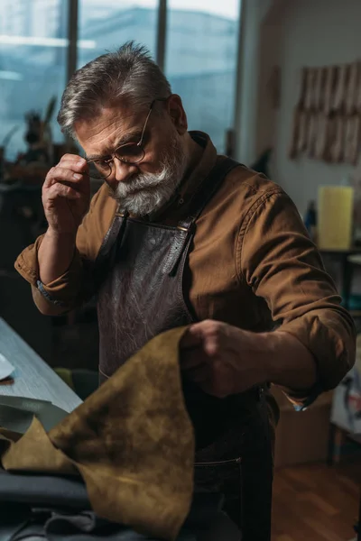 Sapateiro atencioso tocando óculos enquanto olha para pedaço de couro genuíno — Fotografia de Stock