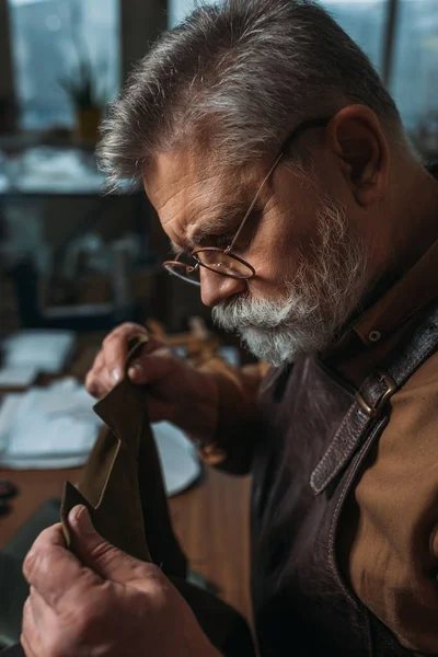 Senior, bearded shoemaker holding piece of genuine leather in workshop — Stock Photo