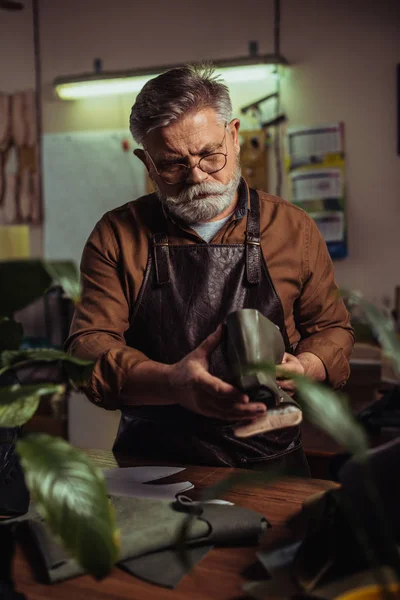 Bearded senior shoemaker holding detail of unfinished shoe in workshop — Stock Photo