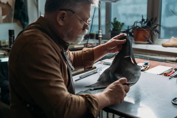 Zapatero senior sosteniendo piezas de cuero genuino en el taller - foto de stock