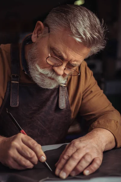 Senior, bärtiger Schuhmacher schneidet echtes Leder mit Messer — Stockfoto
