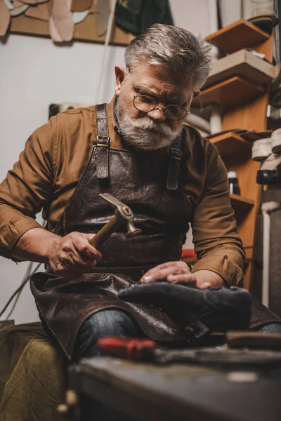 Senior, bärtiger Schuhmacher hält Schuh, während er in der Werkstatt Schuhe repariert — Stockfoto