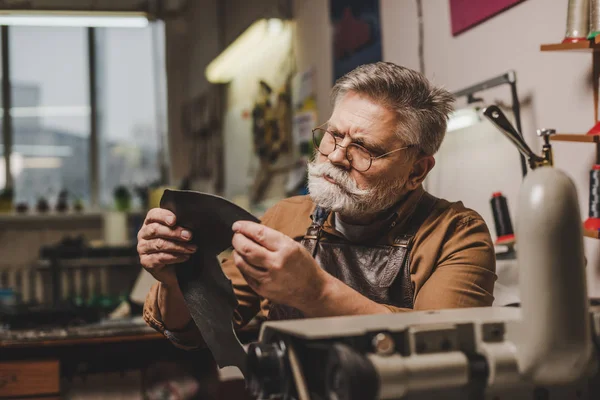 Anciano, zapatero atento que sostiene la pieza de cuero genuino cerca de la máquina de coser - foto de stock