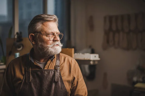Confident, senior cobbler in glasses looking away while standing in workshop — Stock Photo
