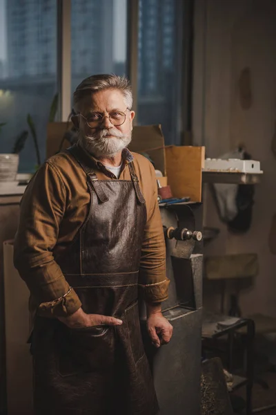 Positiver, bärtiger Schuhmacher in Lederschürze, der in der Werkstatt in die Kamera blickt — Stockfoto