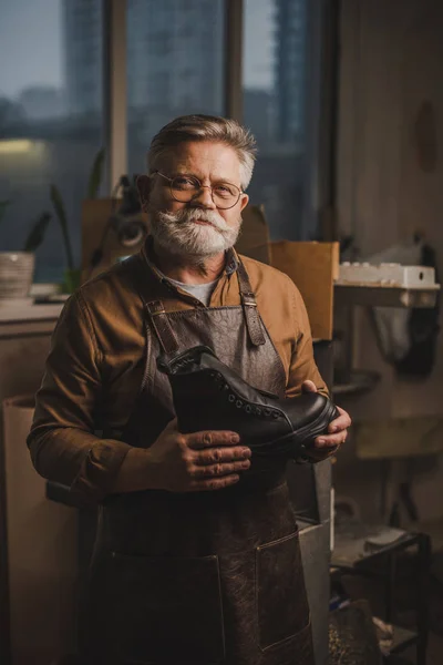 Positive, senior shoemaker smiling at camera while holding leather boot — Stock Photo