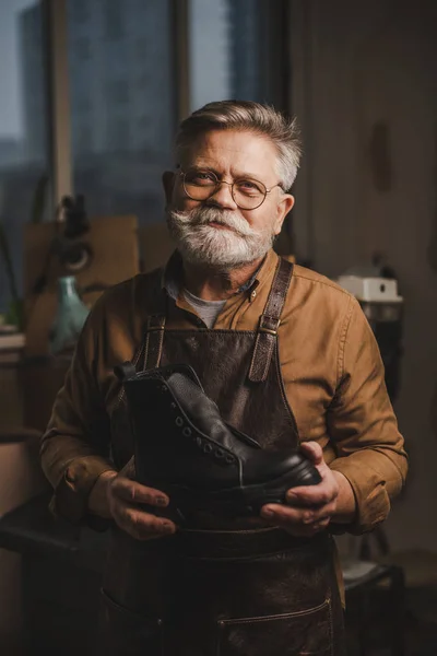 Souriant, barbu cordonnier regardant caméra tout en tenant bottes en cuir — Photo de stock