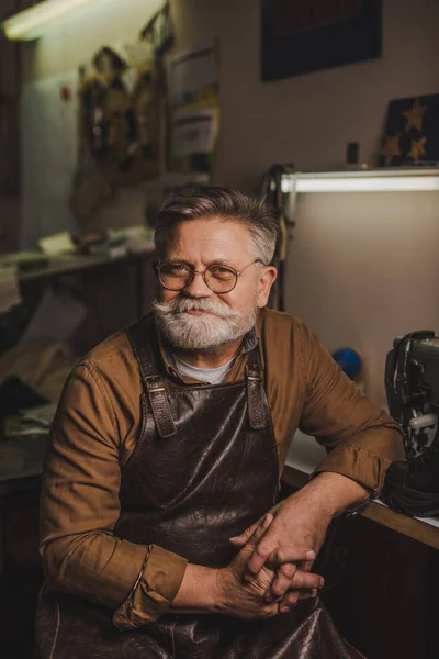 Sonriendo, zapatero senior mirando a la cámara mientras está sentado en el taller - foto de stock