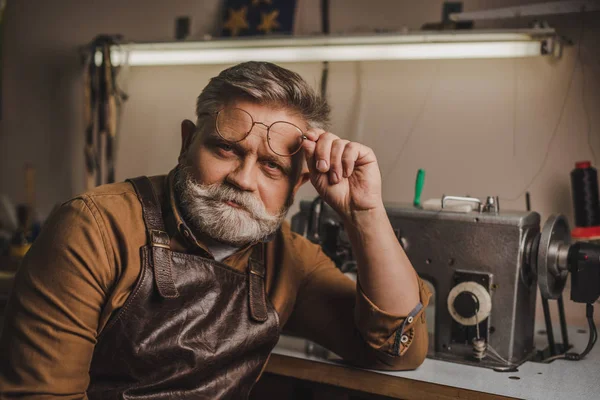 Старший, уверенный в себе сапожник смотрит в камеру, сидя возле швейной машинки в мастерской — стоковое фото