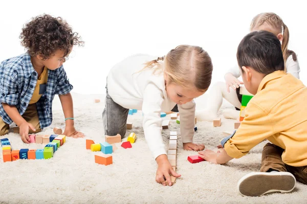 Багатонаціональні діти грають з дерев'яними блоками на килимі, ізольовані на білому — стокове фото
