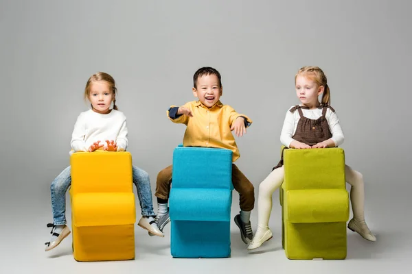 Happy multicultural kids sitting on puzzle chairs, on grey — Stock Photo