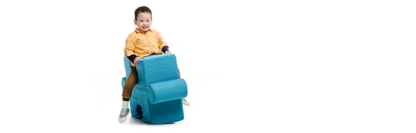 Panoramic shot of happy little asian boy sitting on blue puzzle chair isolated on white — Stock Photo