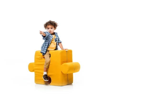 Маленький афроамериканський хлопчик вказує, сидячи на жовтому кріслі-головоломці, ізольований на білому — стокове фото