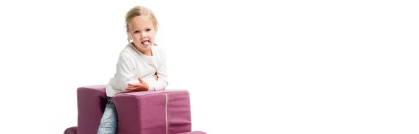 Colpo panoramico di allegro ragazzo attaccare lingua fuori mentre seduto sulla sedia puzzle viola, isolato su bianco — Foto stock
