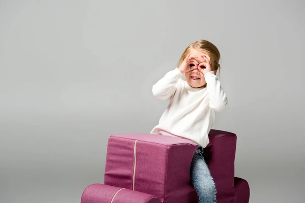 Дитина робить окуляри з рук на фіолетовому кріслі-головоломці, ізольовані на сірому — стокове фото