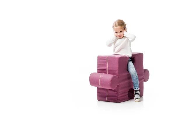 Niño cerrando orejas mientras está sentado en la silla del rompecabezas púrpura, aislado en blanco - foto de stock