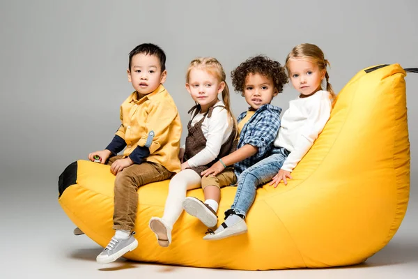 Adorable multicultural kids sitting on bin bag chair on grey — Stock Photo
