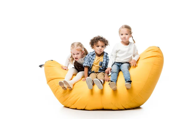 Sad multicultural children sitting on yellow bin bag chair on white — Stock Photo