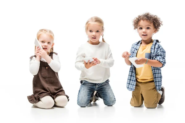 Niños multiétnicos hablando teléfonos inteligentes mientras están sentados en blanco - foto de stock