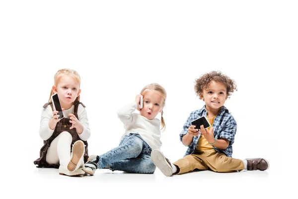 Мультикультурные дети разговаривают на смартфонах, сидя на белом — стоковое фото