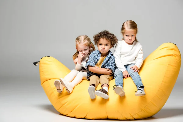 Offended multicultural kids sitting on yellow bin bag chair on grey — Stock Photo
