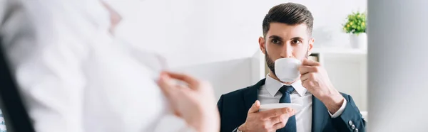 Панорамный снимок бизнесмена, пьющего кофе и смотрящего на секретаря в офисе — стоковое фото