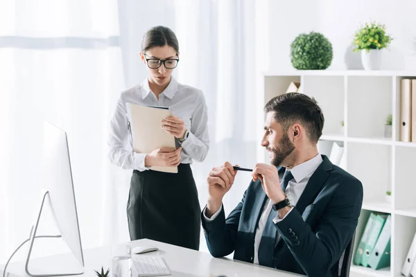 Hombre de negocios guapo sentado en la mesa y mirando a la secretaria en la oficina - foto de stock