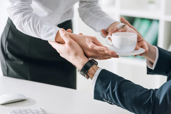 Ausgeschnittene Ansicht von Geschäftsmann, der Sekretärin mit Tasse im Büro die Hand hält — Stockfoto