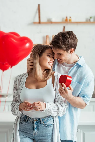 Homme tenant boîte cadeau en forme de coeur et regardant petite amie heureuse avec gros seins — Photo de stock