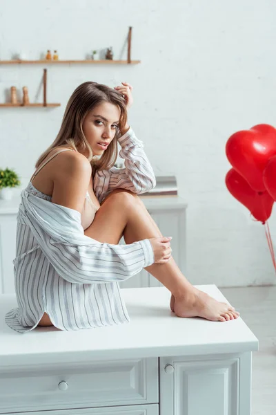 Mulher sexy sentada na mesa perto de balões vermelhos em forma de coração — Fotografia de Stock