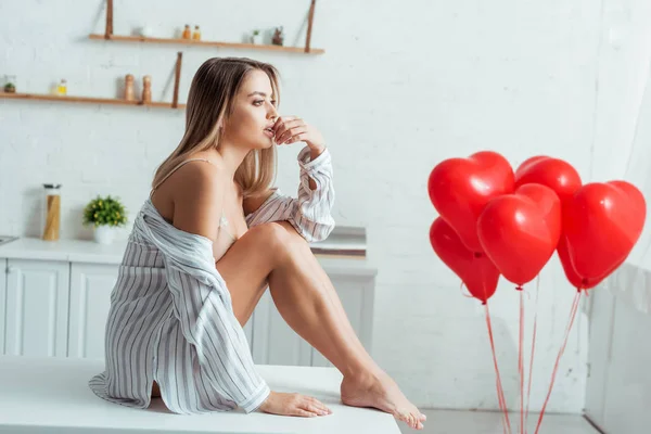 Sexy woman sitting on table and touching lips near red heart-shaped balloons — Stock Photo