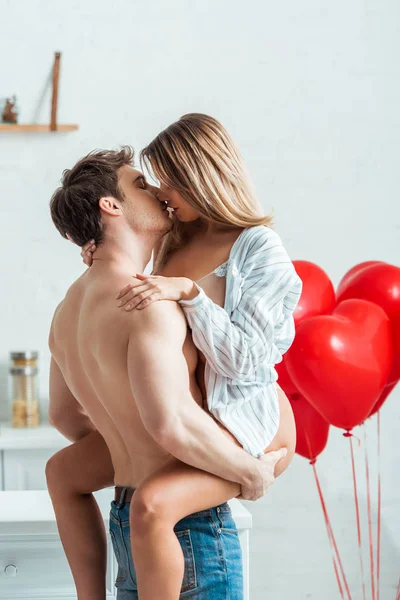 Hombre sin camisa sosteniendo en brazos chica y besándose cerca de globos en forma de corazón - foto de stock