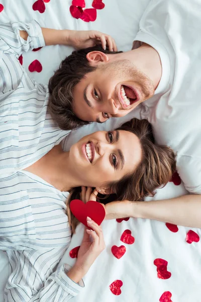 Vista superior de homem alegre e mulher deitada na cama perto de corações vermelhos — Fotografia de Stock