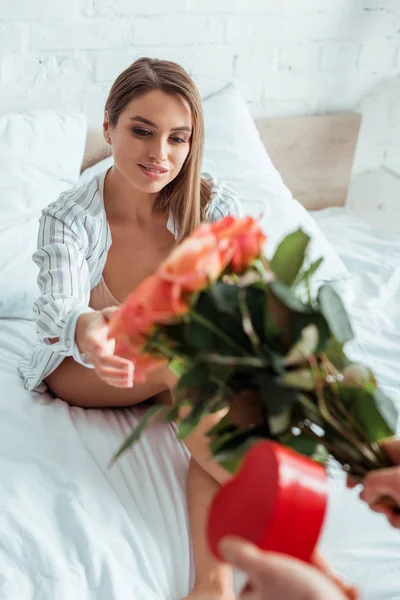 Cropped view of shirtless man giving flowers and heart-shaped gift to cheerful woman in bedroom — Stock Photo