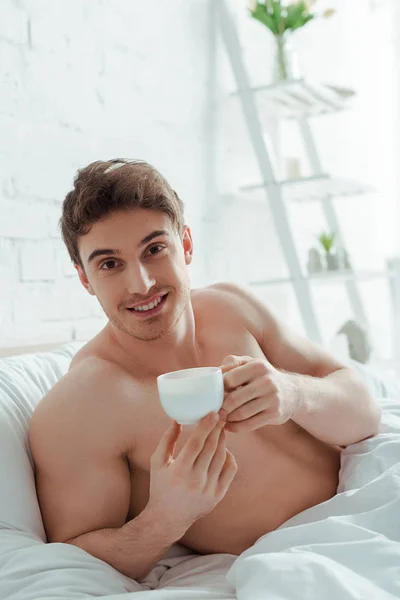 Happy and shirtless man holding cup of coffee in bed — Stock Photo
