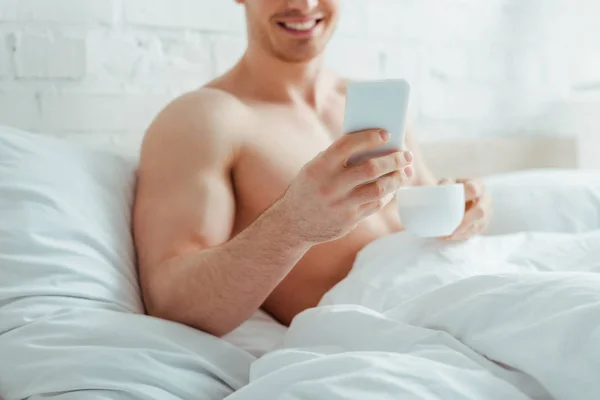 Vista recortada de hombre feliz sin camisa sosteniendo la taza de café mientras usa el teléfono inteligente en la cama - foto de stock