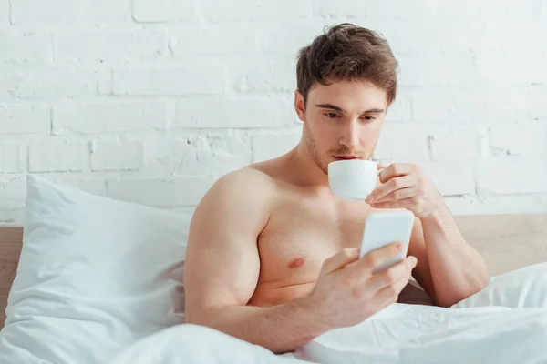 Hombre sin camisa guapo sosteniendo la taza de café mientras usa el teléfono inteligente en la cama - foto de stock