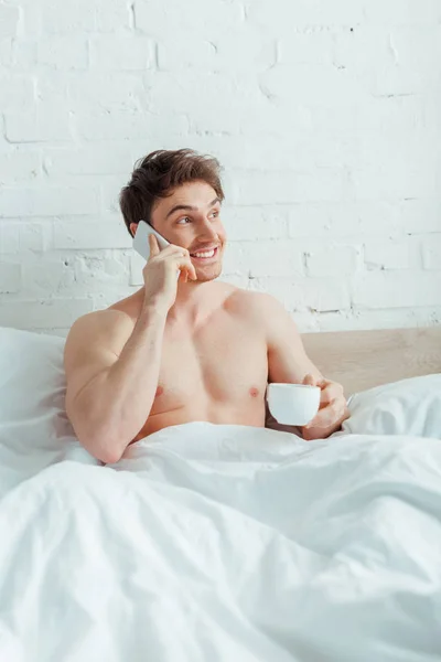 Hombre feliz y sin camisa sosteniendo la taza de café mientras habla en el teléfono inteligente en la cama - foto de stock